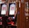 A pair of casino slot machines, each with detailed screens displaying various game outcomes and jackpot amounts. The chairs in front of the slot machines are dark and cushioned for comfort. The background features a hanging curtain made of golden ropes and other casino decorations are visible.